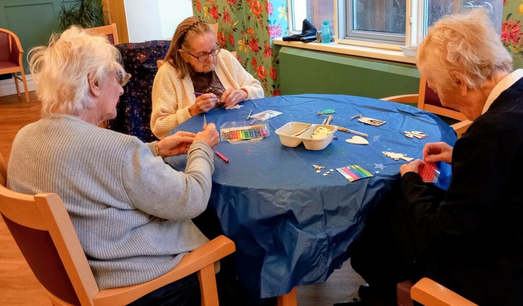 Three elderly women sit around a blue tablecloth, making white ornaments and coloured threads for the St Hildeburgh’s Festival tree in a brightly lit room with floral wallpaper. Each piece is crafted with care.