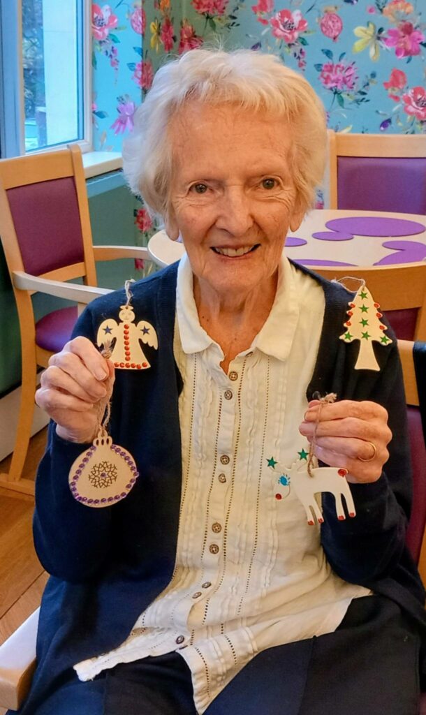 An elderly woman sits in a decorated room, smiling and holding handmade Christmas decorations—a tree, angel, heart, and reindeer—all crafted with care for St Hildeburgh’s Festival.