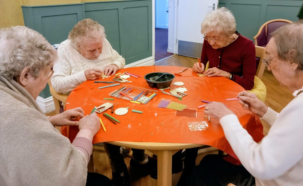 Four elderly women sit around a table covered with craft materials, engrossed in arts and crafts activities in a bright room—every handmade creation carefully crafted for St Hildeburgh’s Festival.