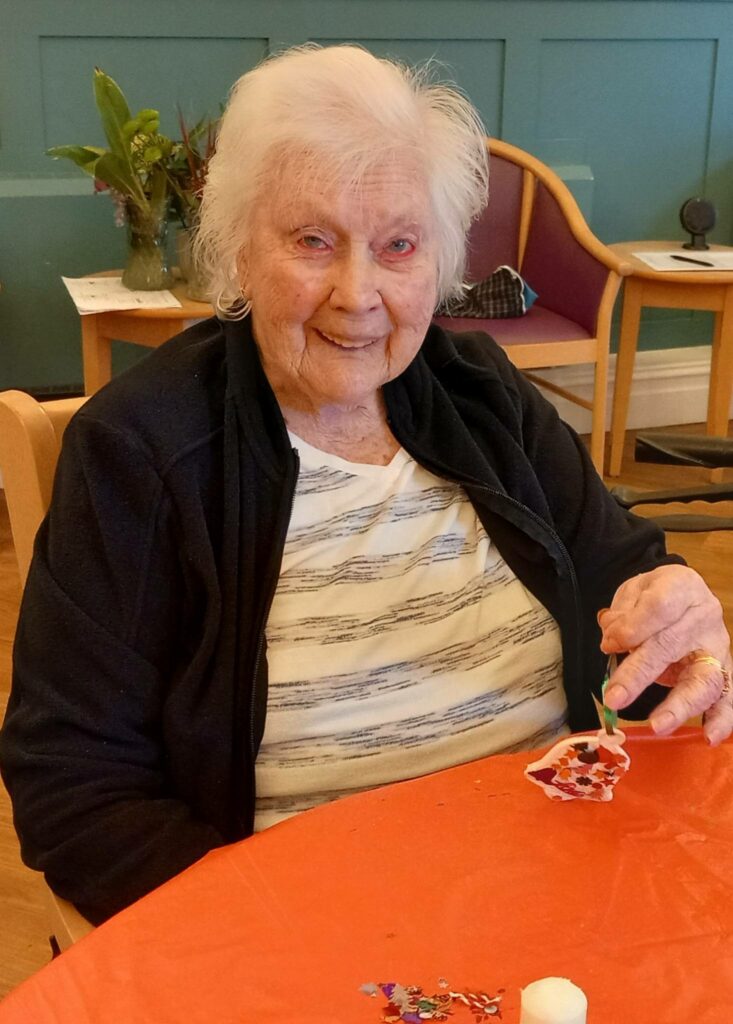 Elderly woman seated at a table, carefully holding a decorated ornament and smiling at the camera, celebrating St Hildeburgh’s Festival.