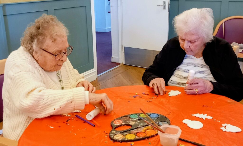 Two elderly women sit at a table doing arts and crafts with paint, glue, and paper shapes on a bright orange tablecloth during St Hildeburgh’s Festival, creating a colourful tree design together.