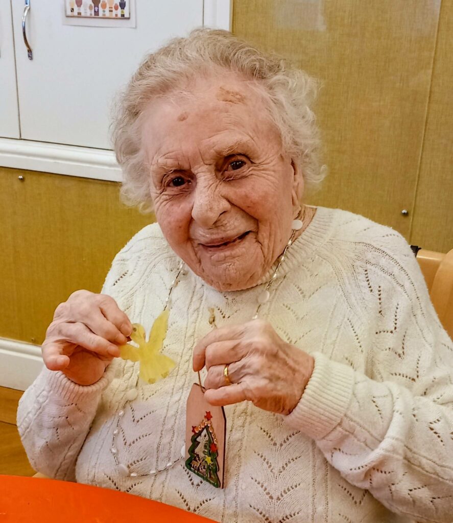 Elderly woman in a white jumper, smiling as she holds a yellow paper craft and a carefully made Christmas tree ornament at St Hildeburgh’s Festival.