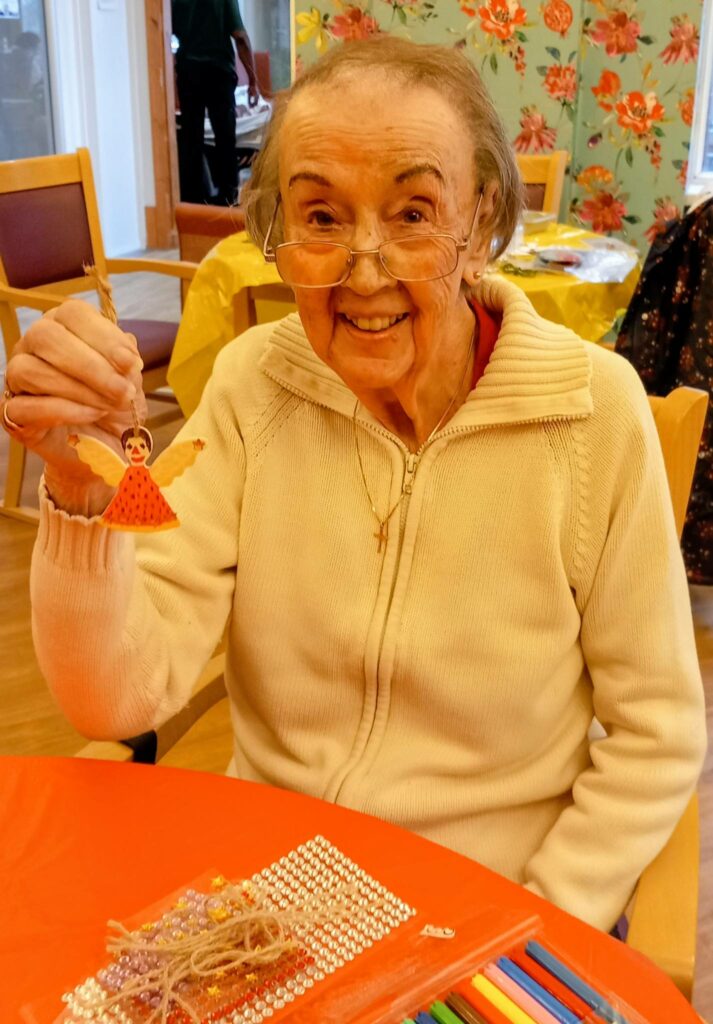 Elderly woman wearing glasses smiles and holds up a carefully made handmade craft doll, seated at a table with art materials and decorations at St Hildeburgh’s Festival.