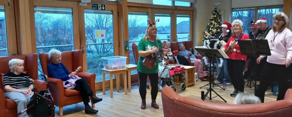 A small group of musicians in festive attire perform for seated elderly ladies in a decorated room with a Christmas tree.