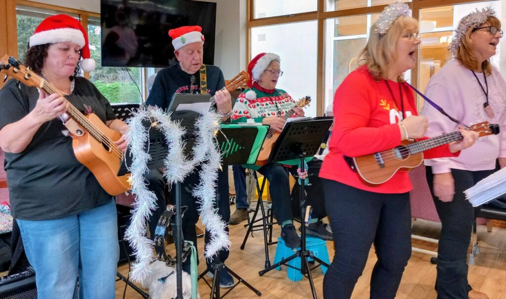Five adults dressed in festive clothing, including Father Christmas hats and Christmas jumpers, play musical instruments and sing together indoors, likely at a holiday gathering.