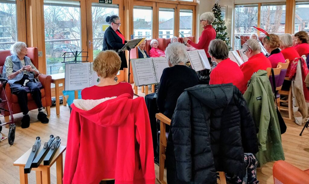 A group of older adults in red clothing, known as The Beacon Belles, play music with music stands indoors on a festive afternoon, while others listen, seated near large windows overlooking a wintry scene.