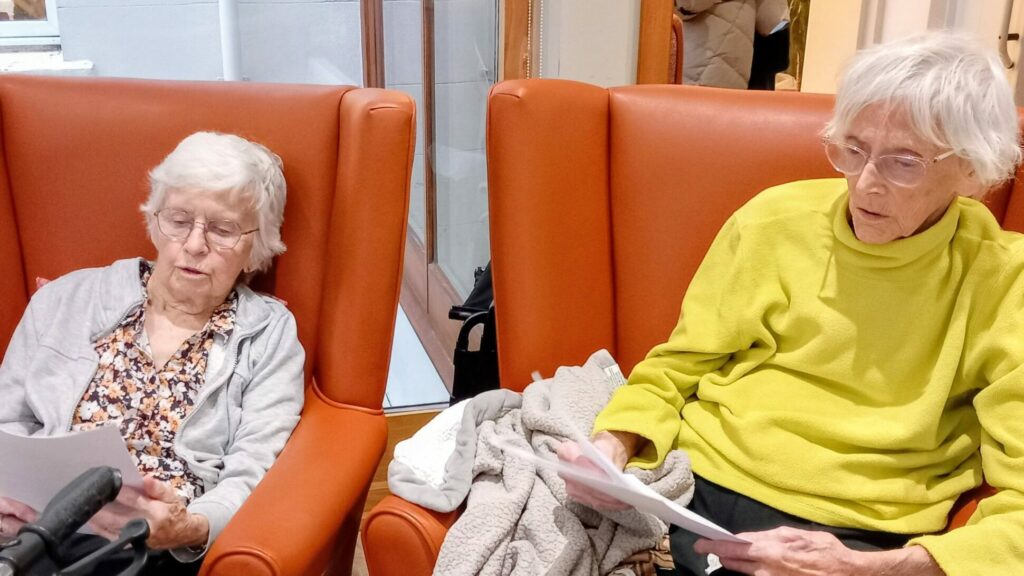 Two elderly women sit in orange armchairs, each holding a sheet of paper and reading, with a beige blanket draped over one of the chairs.