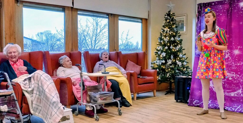 A performer dressed in a colourful clown costume entertains four elderly women seated in armchairs beside a Christmas tree in a care home lounge.