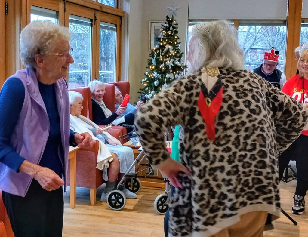 Elderly people gather in a decorated room with a Christmas tree, chatting and laughing; some are seated while two women stand in the foreground.