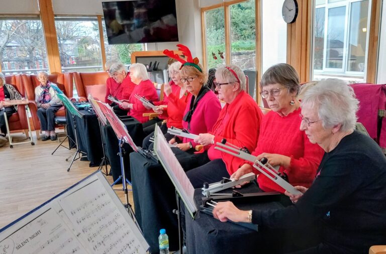 The Beacon Belles, a group of elderly women dressed in red, sit in a semi-circle with chimes and music stands, performing on a festive afternoon; one lady playfully wears antlers.