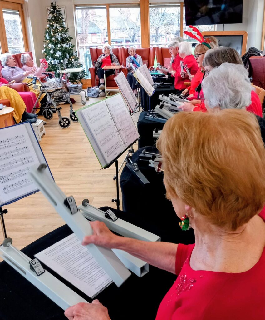 The Beacon Belles, elderly women in red tops, play hand chimes from sheet music while others listen near a decorated Christmas tree during this festive afternoon in a bright room.