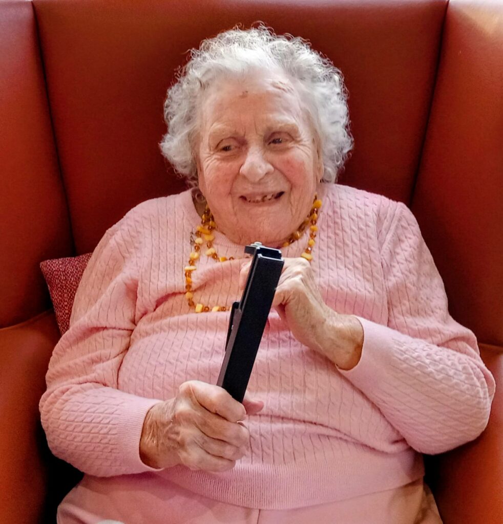 Elderly woman with curly grey hair sits in a red armchair, smiling and holding a remote, enjoying a festive afternoon as one of the cheerful Beacon Belles.