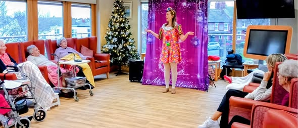 A performer in a colourful dress entertains a group of elderly people seated in a lounge with a Christmas tree and festive decorations.