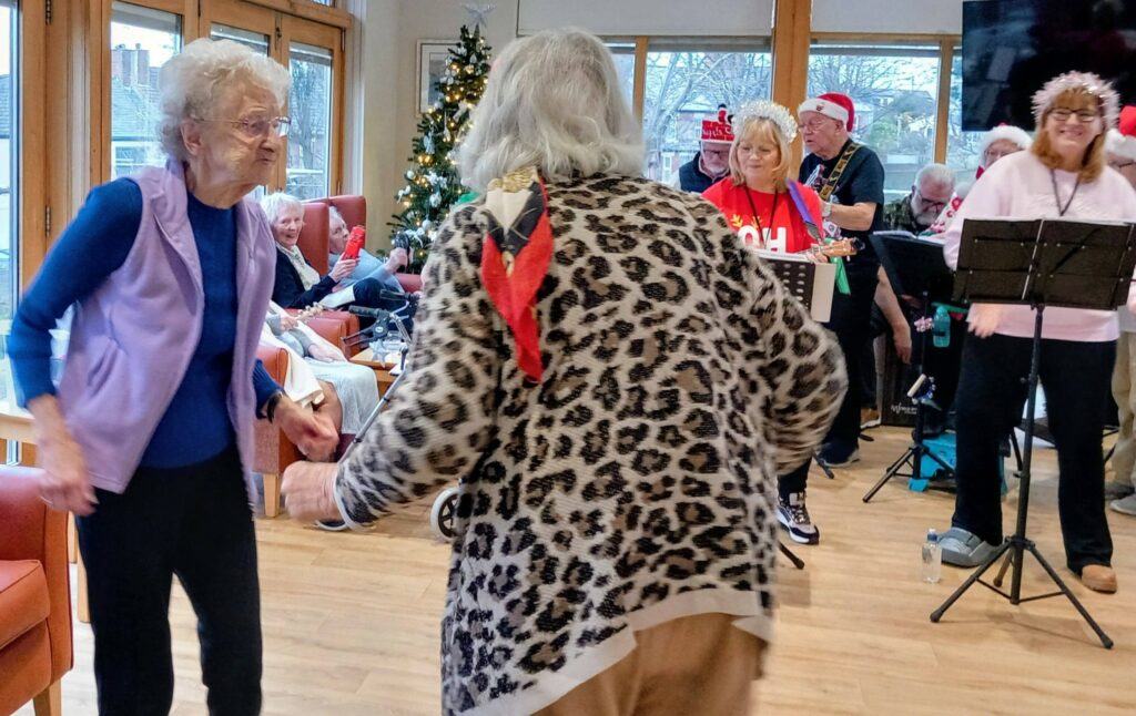 Two elderly women dance together in a brightly lit room as a small band plays music; people sit and watch nearby, next to a decorated Christmas tree.