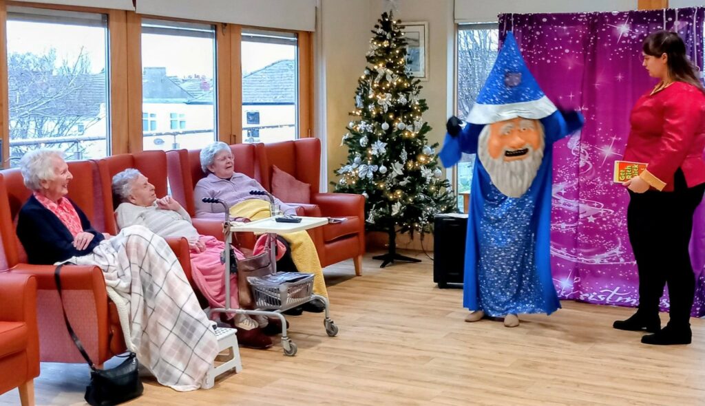 Four elderly women watch a performer in a blue wizard costume and a woman holding a book in a festive room with a Christmas tree and purple backdrop.