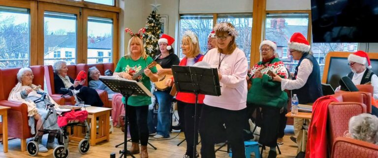 A group of people in festive attire and Father Christmas hats perform music for seated elderly residents in a decorated lounge, with a Christmas tree in the background.