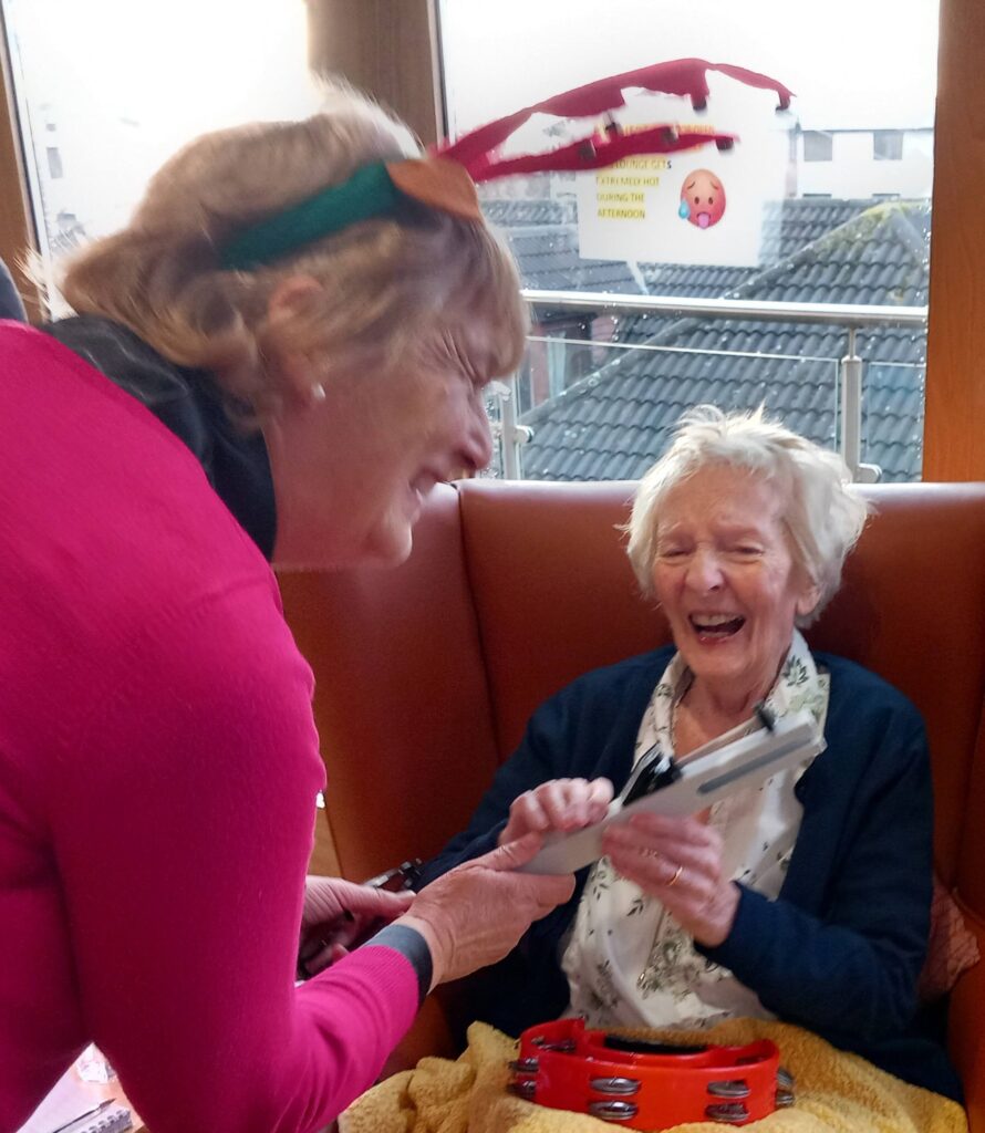 Two elderly women smile and laugh together on a festive afternoon; one wears reindeer antlers, and the other holds a tambourine while sitting on a sofa, enjoying the company of fellow Beacon Belles.