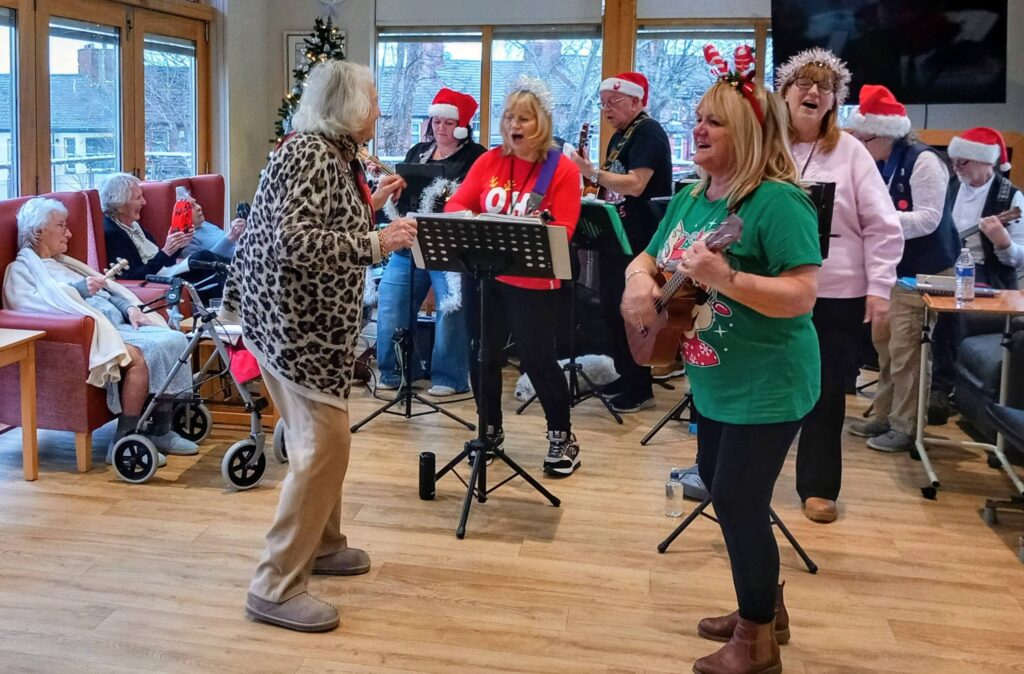 A group of people in festive hats perform music and sing in a care home lounge as residents watch, with one resident dancing at the front.