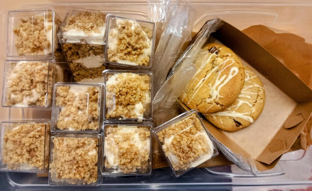 Plastic containers filled with crumb-topped puddings are gathered next to a cardboard box containing several biscuits drizzled with white icing.