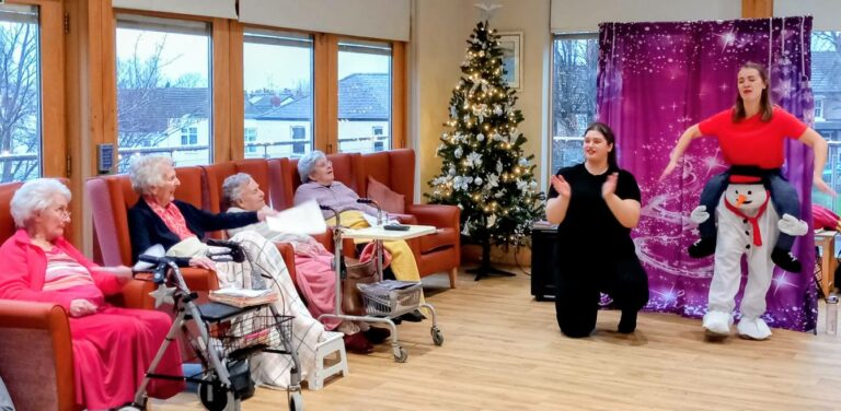 A group of elderly women sit on chairs watching two performers, one appearing to ride a snowman, in a room decorated with a Christmas tree and festive decorations.