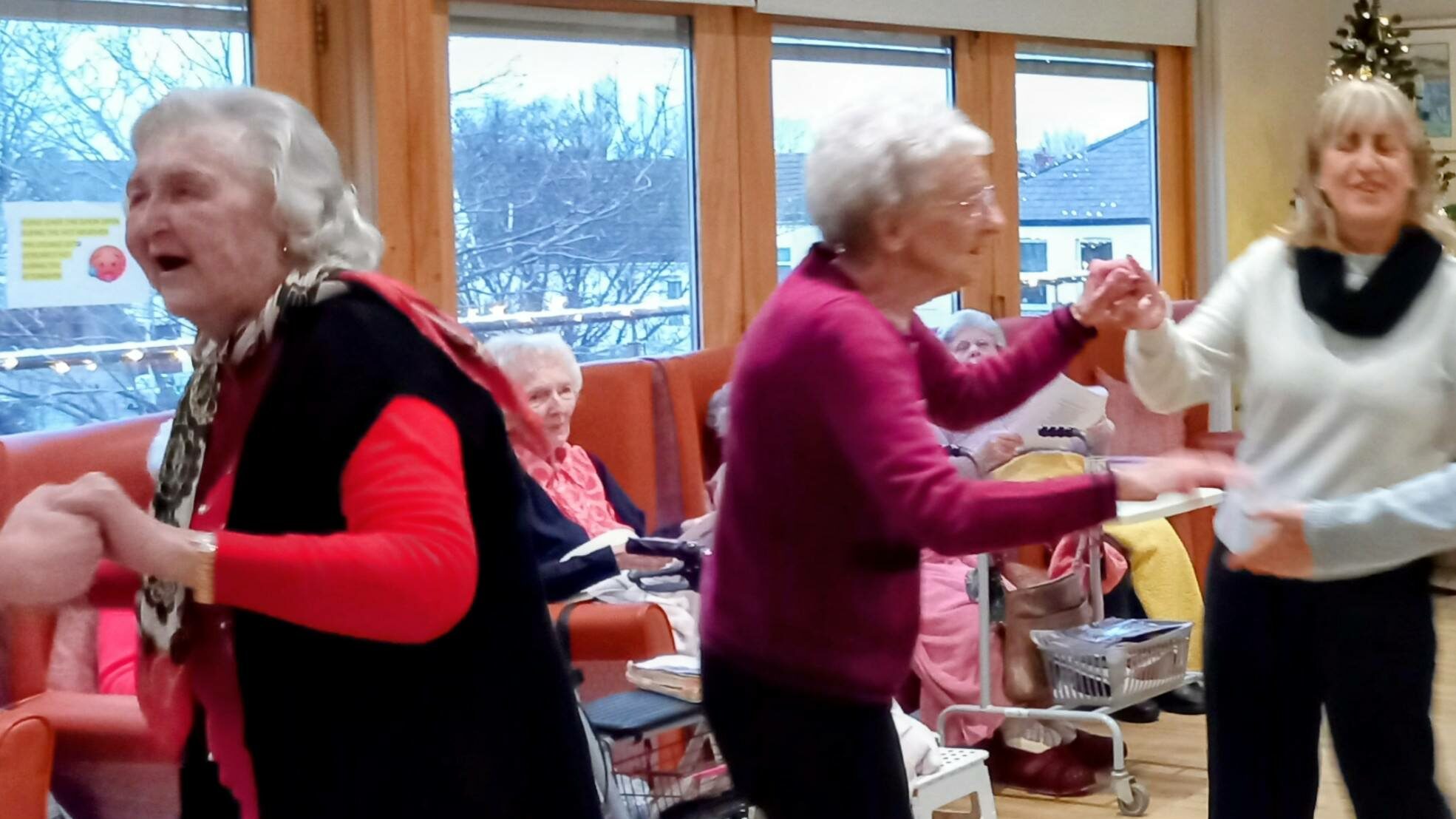 Three elderly women are dancing together in a communal room, while other pensioners sit and watch in the background.