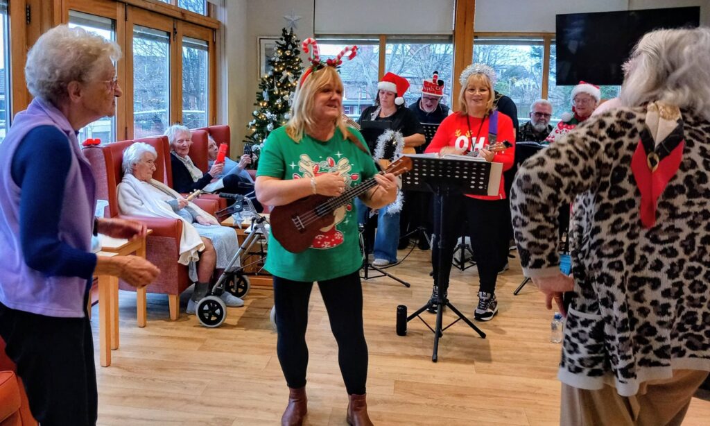 A group of elderly people enjoy a festive music performance with singers and musicians in seasonal attire at a care home.