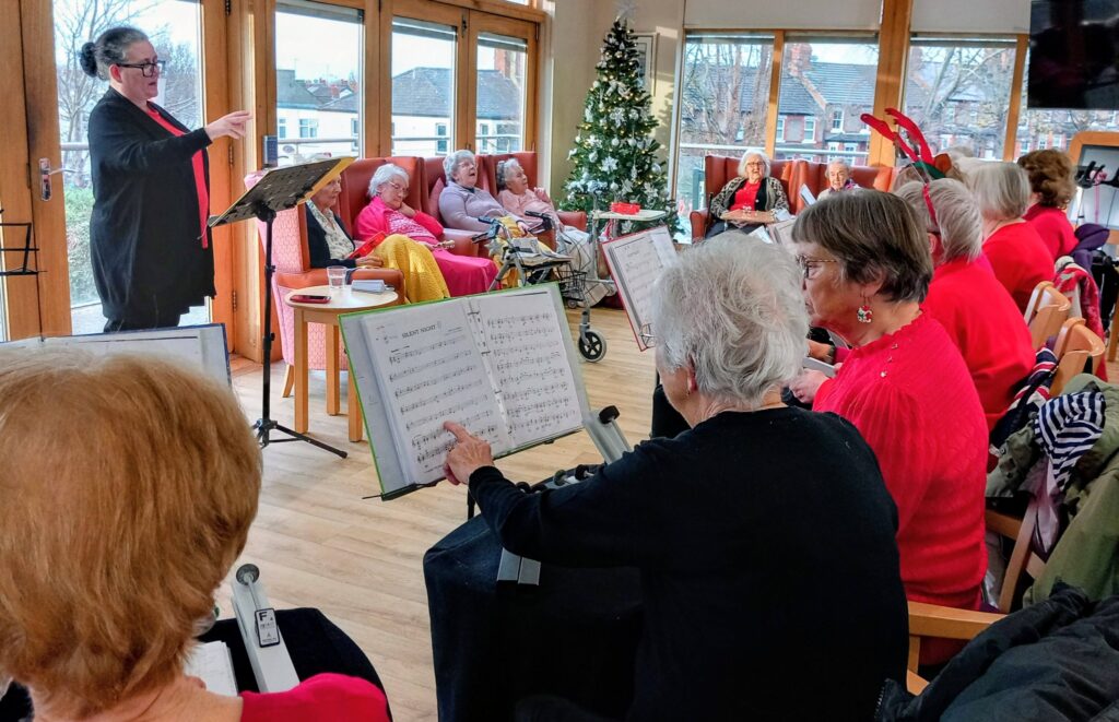 The Beacon Belles perform for an audience of elderly people in a bright room with a Christmas tree, creating a festive afternoon as the conductor leads and sheet music is visible in the foreground.