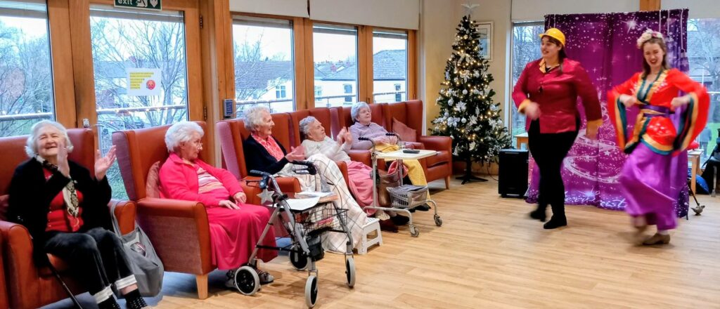 A group of elderly women sit in armchairs, watching and applauding as two performers in colourful costumes dance in a decorated room with a Christmas tree.