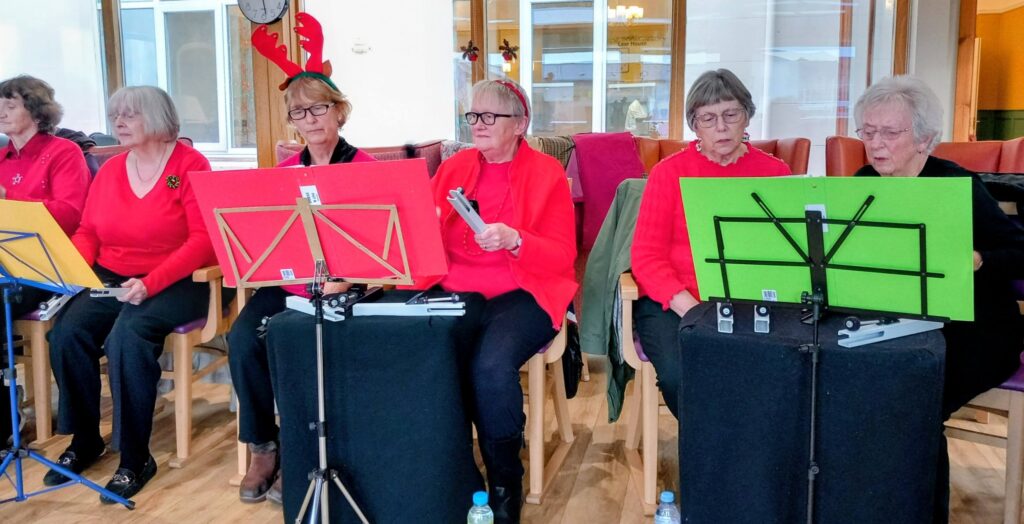 Five elderly women, the Beacon Belles, sit on chairs with music stands and red tops. One is wearing reindeer antlers, prepared for a festive afternoon rehearsal or performance indoors.