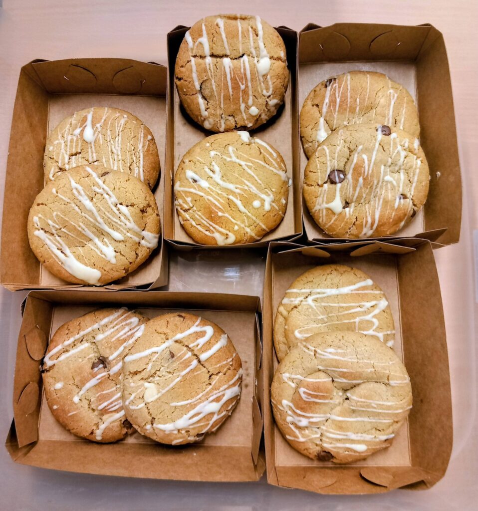 Six cardboard trays, each holding two large biscuits topped with a white icing drizzle, are set out in two rows on a light-coloured surface.