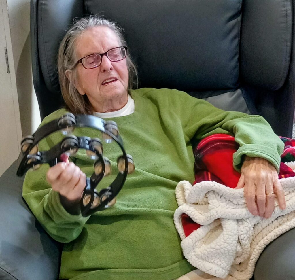 An elderly woman sits in an armchair, holding a tambourine in one hand and a blanket in the other. She is wearing spectacles and a green jumper.
