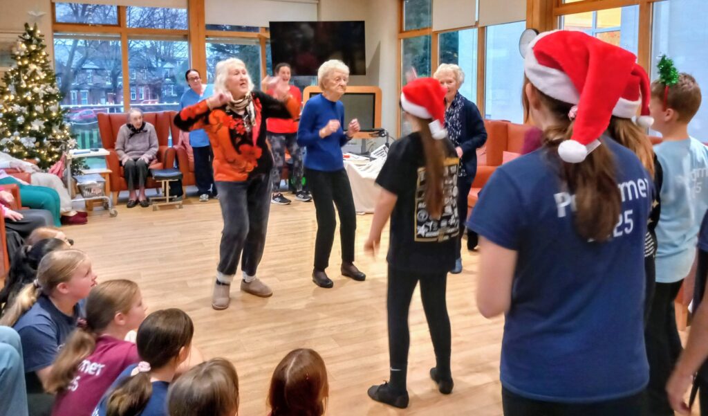 Elderly women dance enthusiastically as children in Santa hats and adults watch in a festively decorated room with a Christmas tree.