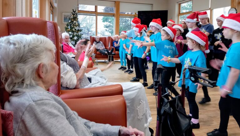 A group of children in Santa hats perform for elderly residents seated in the lounge of a care home, with a Christmas tree visible in the background.