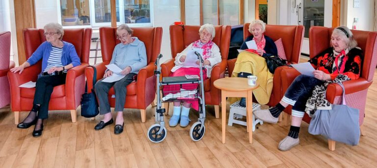 Five elderly women sit in armchairs holding papers, with a walking frame and a table with a mug in front of them, in a brightly lit room with wooden flooring.