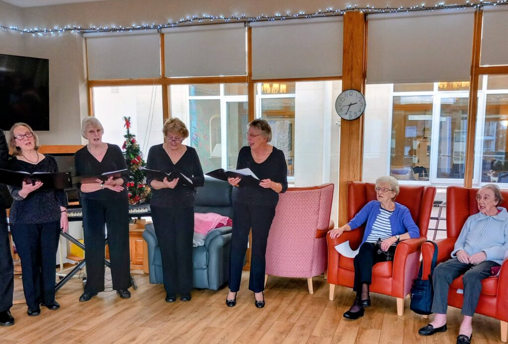 Five women stand singing before a Christmas tree, as three elderly women sit watching in a brightly lit room furnished with armchairs and a wall clock.
