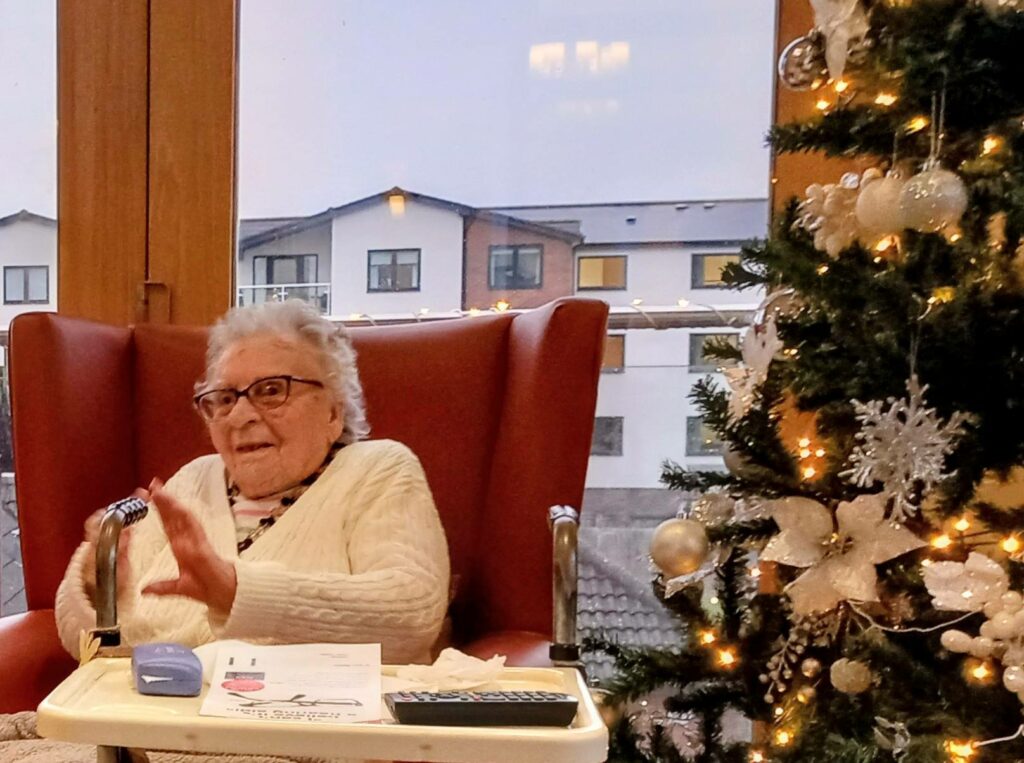 An elderly woman sits in an armchair by a window beside a decorated Christmas tree, with papers and a remote on a tray in front of her.