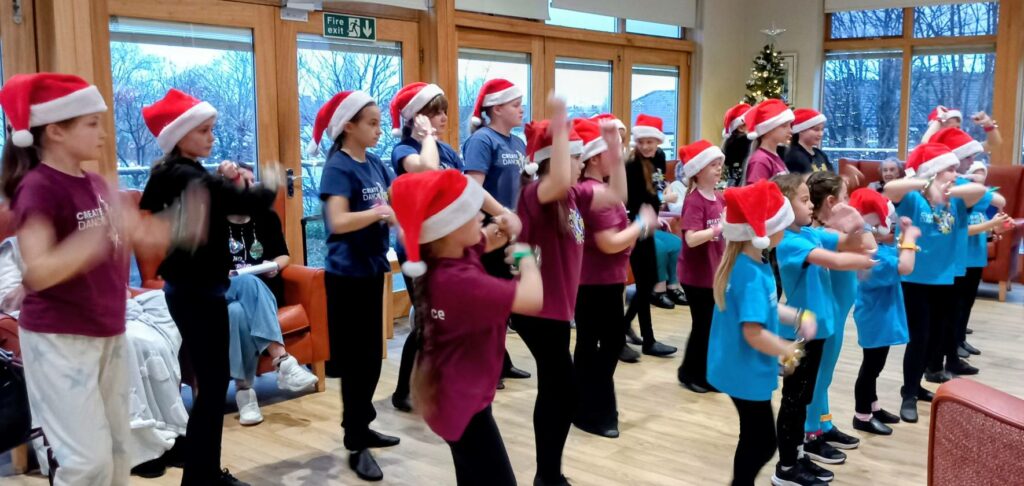 A group of children and teenagers wearing Santa hats perform a choreographed dance indoors, with an audience seated in the background and a Christmas tree visible.