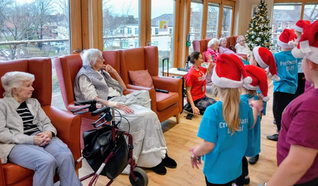 Children in festive hats perform for elderly residents seated in a care home lounge adorned with a Christmas tree.