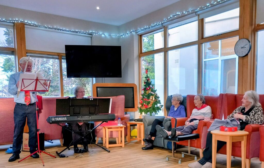 A man plays the flute and a woman plays the keyboard for four elderly women seated in a lounge adorned with Christmas lights and a tree.