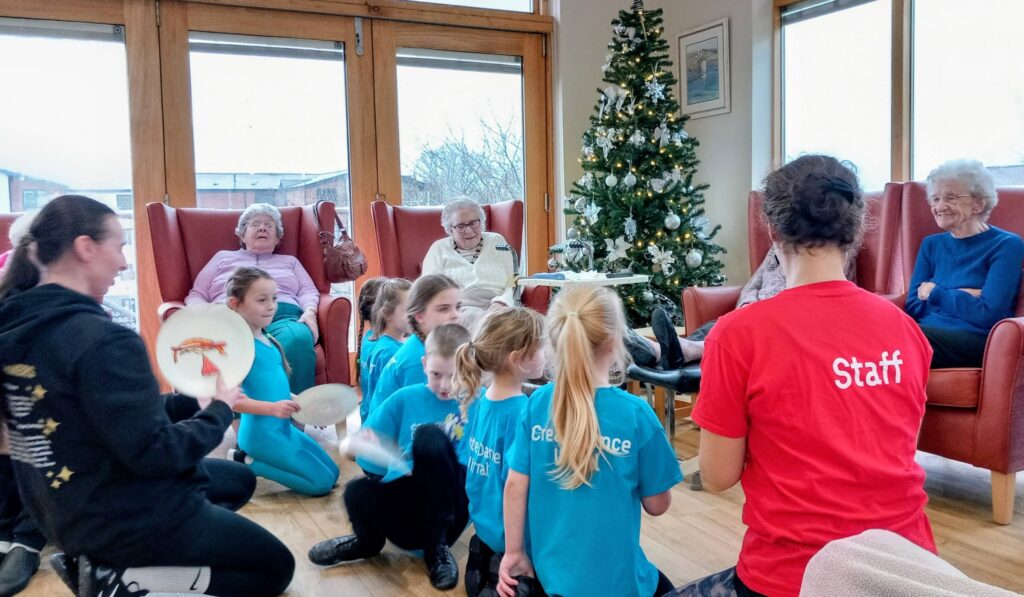 Children perform for elderly ladies seated by a Christmas tree in a care home; staff members are present, and the children are holding tambourines.