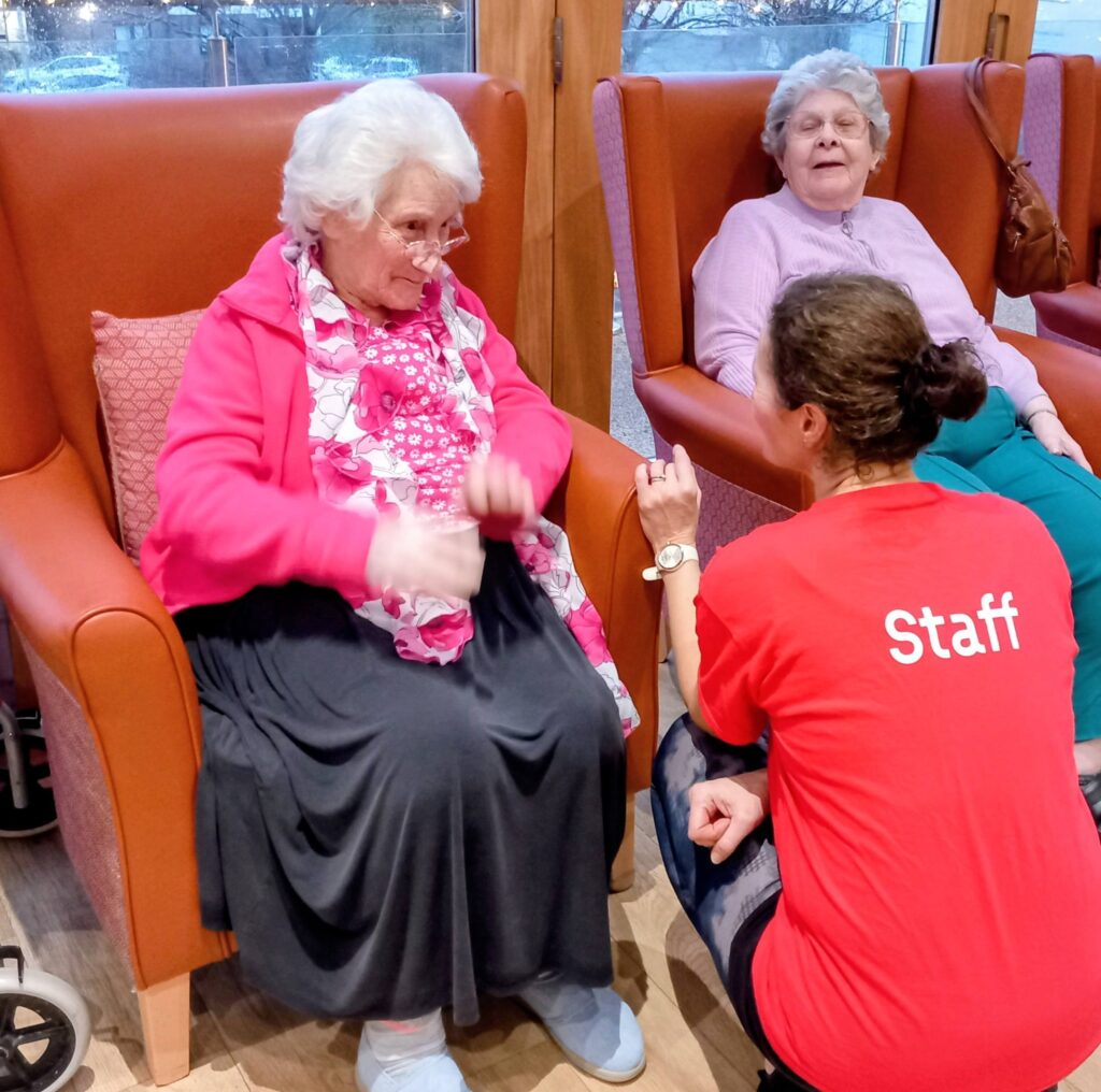 A staff member in a red shirt kneels and chats with two elderly women seated in armchairs in a bright room.