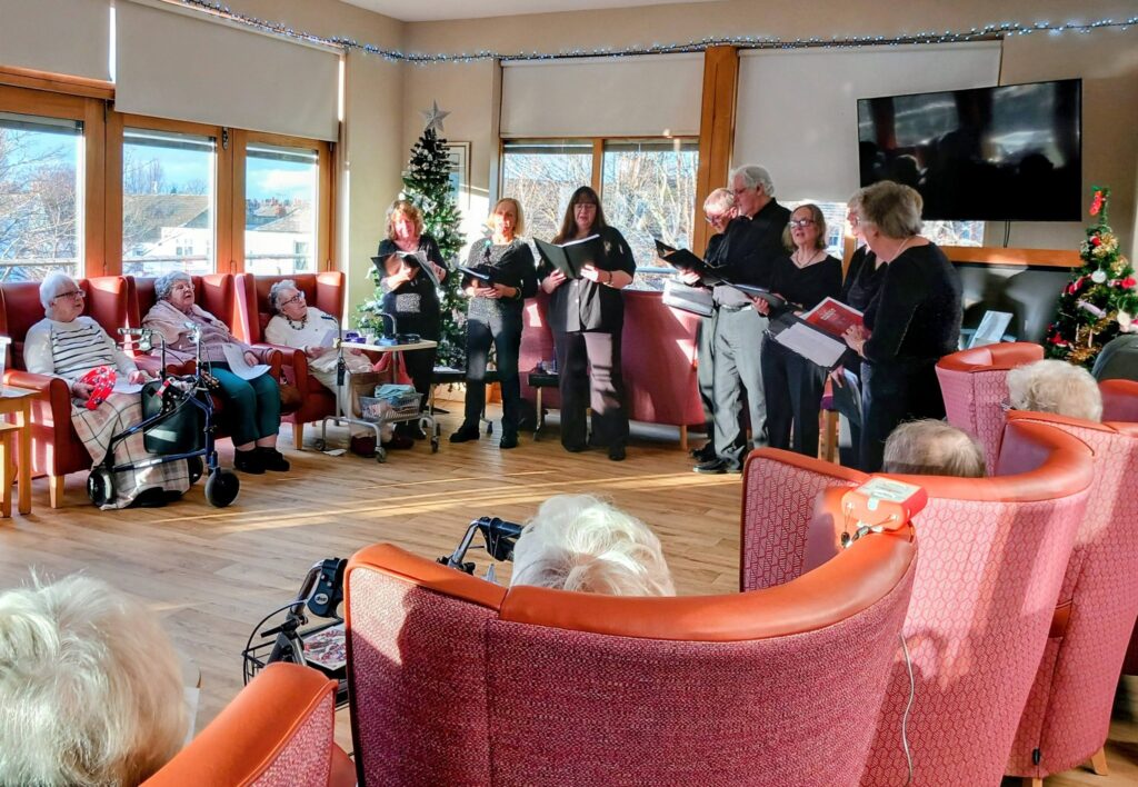 A group of people stand singing from songbooks in a brightly lit room adorned with festive decorations, while elderly residents sit and watch from armchairs.