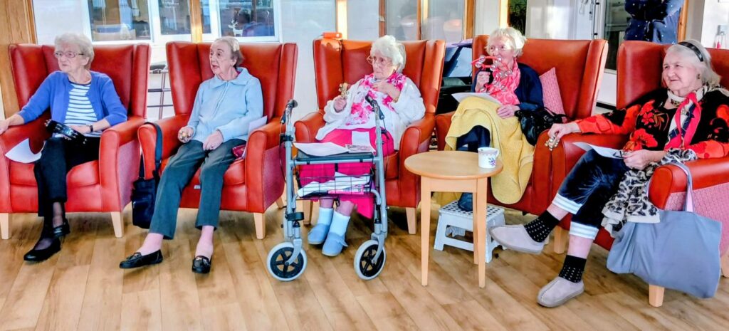 Five elderly women sit indoors on a row of orange chairs; some hold papers or drinks, with a walking frame and a mug visible in front of them.