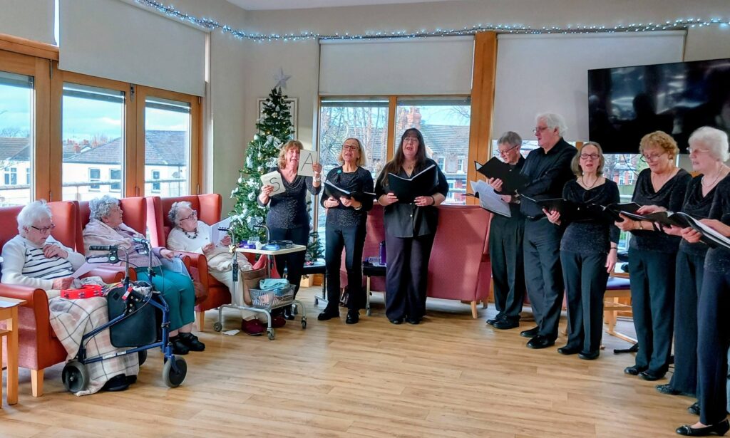 A choir sings to elderly residents seated in a lounge adorned with a Christmas tree and fairy lights.
