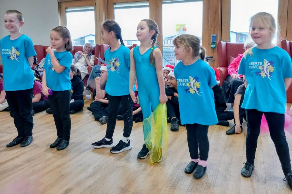 Six young children wearing blue Create Dance shirts stand in a line indoors, getting ready to perform, while an audience is seated in the background.