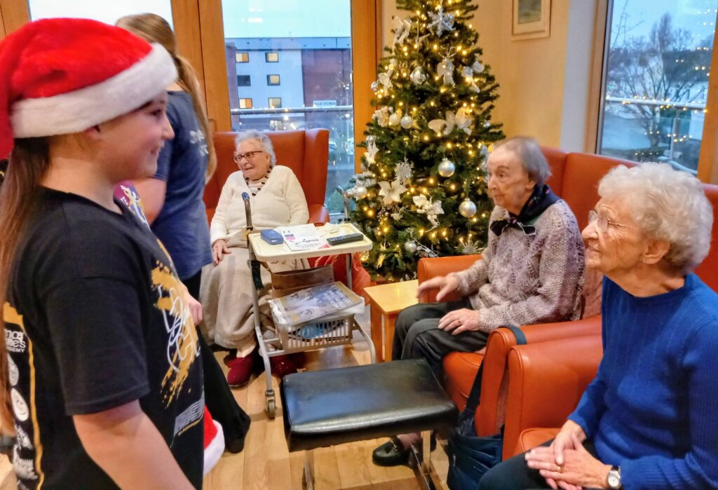 A young person wearing a Santa hat smiles and chats with three elderly women seated beside a decorated Christmas tree in a communal lounge.