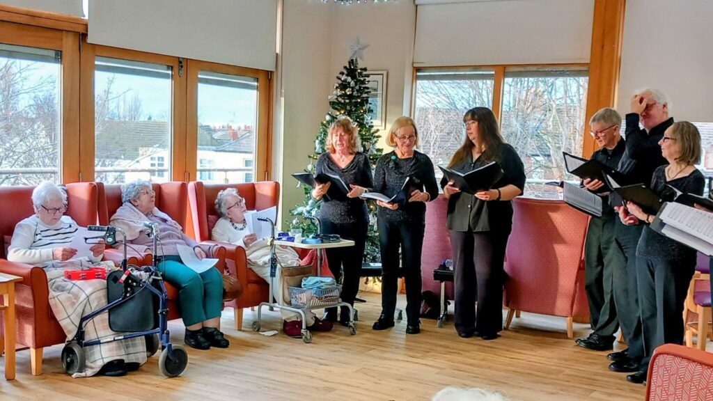 A group of people sing from songbooks to elderly residents seated in a care home lounge adorned with a Christmas tree.