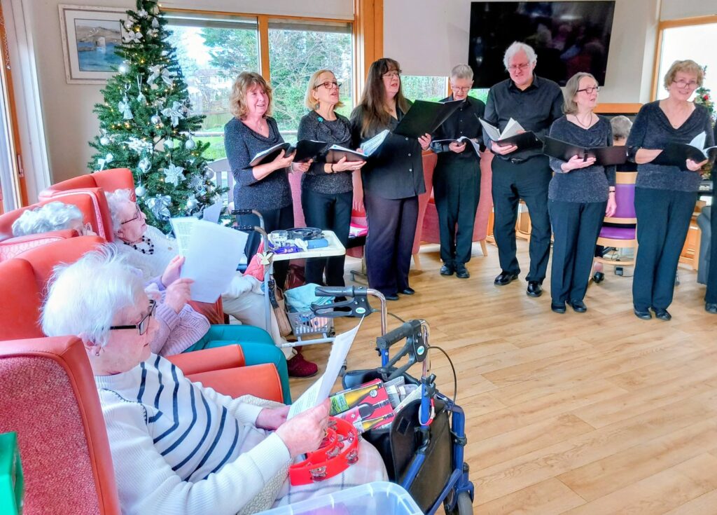 A choir sings to a group of elderly people seated in a brightly lit room adorned with a Christmas tree. Some residents hold song sheets or use walking frames.
