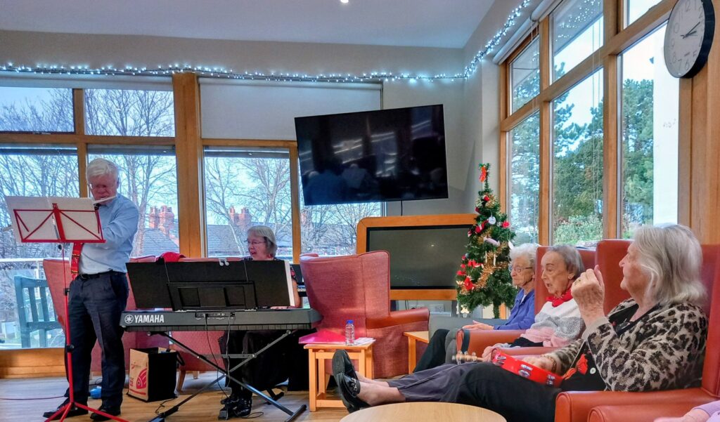 A small group of elderly people sit in a lounge watching a man play music on a keyboard, with festive lights and a Christmas tree in the background.
