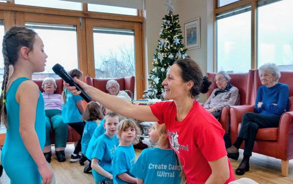 A woman in a red top holds a microphone for a young girl in a blue leotard as other children and elderly people look on in a room decorated with a Christmas tree.
